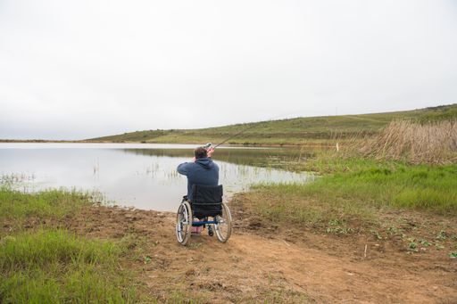 The man in a wheelchair is fishing near the lake.
