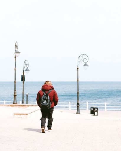A person in a red jacket walks along a seaside promenade with vintage street lamps and a clear blue ocean view.