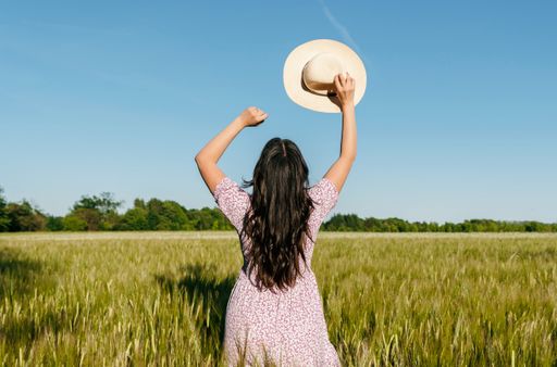 Lifestyle image of beautiful woman with long hair, walking in field of wheat or barley, holding hat with raised arms.