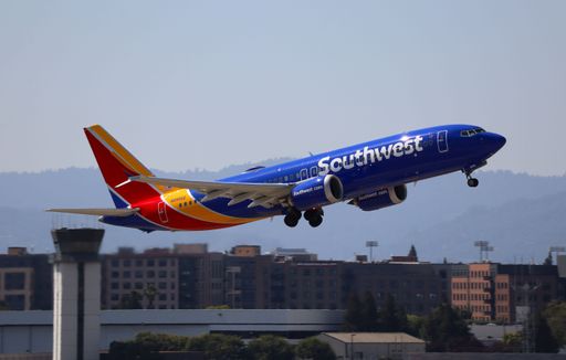 A Southwest Airlines plane takes off from an airport with urban buildings and mountains in the background.
