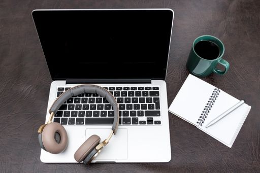 A closeup of a laptop on a desk with headphones on the keyboard, notebook, pen, coffee on the side.