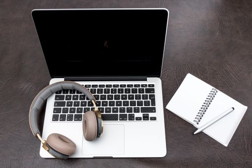 A closeup of a laptop on a desk with headphones on the keyboard, a notebook, and a pen on the side.