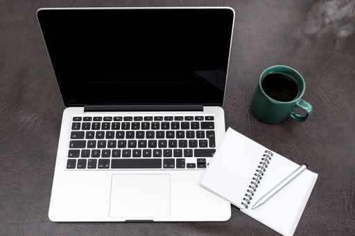 A closeup of a laptop on a desk with a notebook, pen, and cup of coffee on the side.