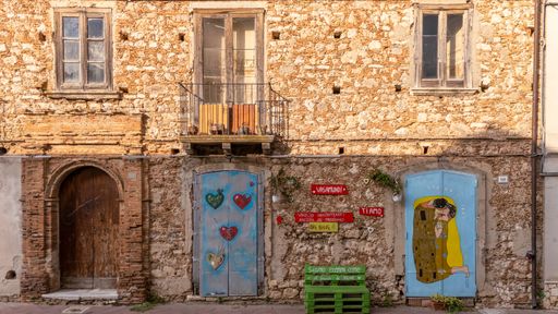 Painted facade of an abandoned house in Mileto with blue doors, hearts, and a mural of a kissing couple.