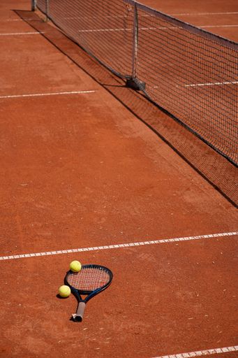 A tennis racket and balls on a clay court under bright sunlight, showcasing the sport's essentials.