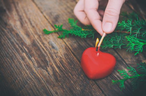 A hand lighting a heart-shaped candle on a wooden table with spruce branches