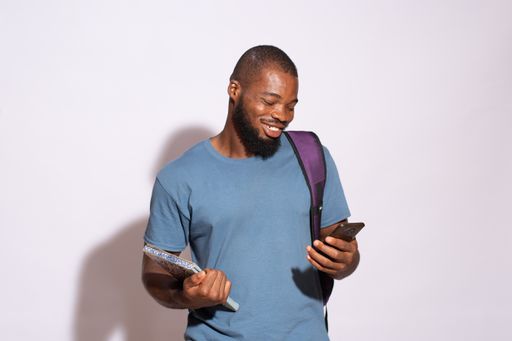 A smiling African male student looking at his phone while standing against a white wall