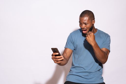 A surprised African male student looking at his phone while standing against a white wall