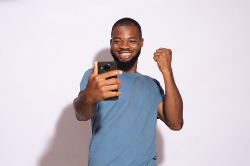 An excited African male student looking at his phone while standing against a white wall