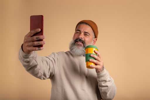 A handsome Spanish man with a white beard and brown hat taking selphie with a red smartphone