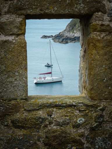 A vertical shot of a sailboat seen through a small window in a textured stone wall in France