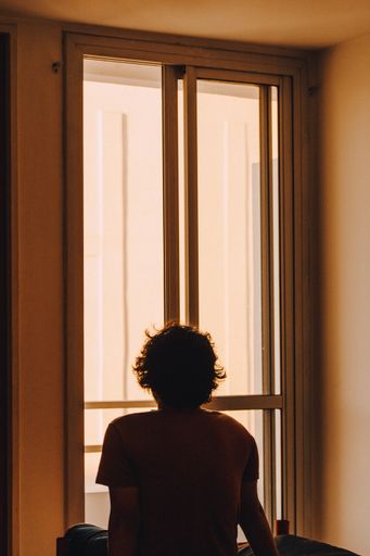 A short curly-haired person sitting on a bed and looking out of the window on a sunny day