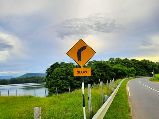 A yellow right curve road sign on a pole near the lake under a blue cloudy sky