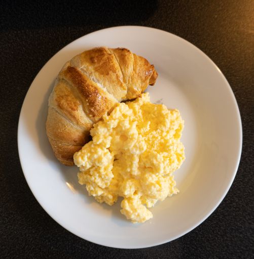 A top view of scrambled eggs and a croissant on a white plate on the table