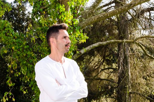 A Caucasian man in profile with white shirt and mustache with crossed arms on background of trees.