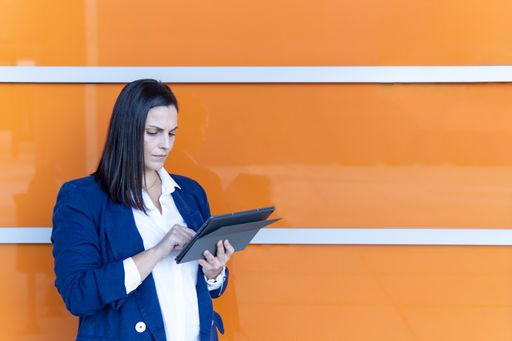 Beautiful Caucasian mature businesswoman using a digital tablet, working in the street outside of an office building, orange background, copy space