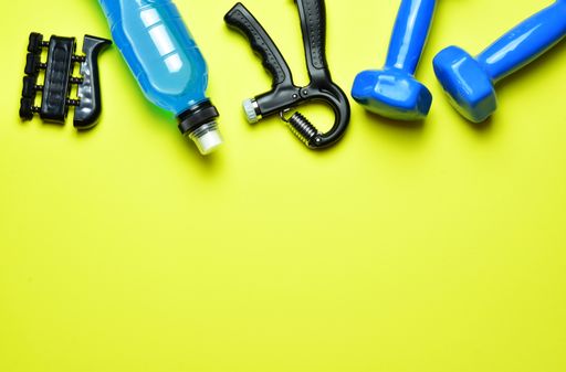 A top view of fitness tools. The water bottle, hand and finger grips, towel, and dumbbells isolated on yellow background