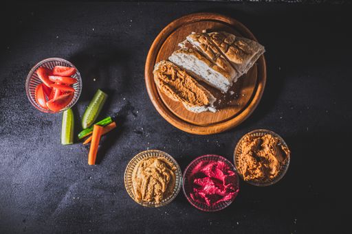 A top view of sliced bread, vegetables and bowls of homemade hummus on a black table