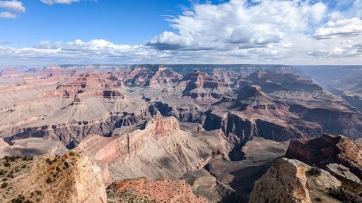 Panoramic view of the Grand Canyon under a bright blue sky with scattered clouds, showcasing its vast and rugged beauty.