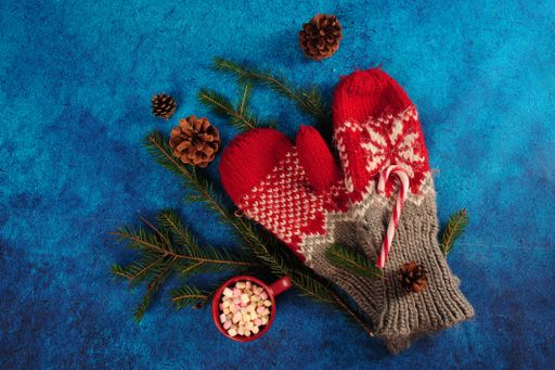 A top view of a Knitted grey and red mittens, marshmallows, pinecones, pine twigs and cup with tiny marshmallows on a blue surface.