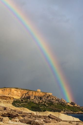 Dramatic rainbow against dark stormy sky, over watchtower on coastal cliff, fields shelving down towards sea, and limestone rocky shore. Malta,