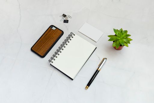 A flat lay of a blank notepad, pen, phone, clips, paper cards, and a plant on a white table