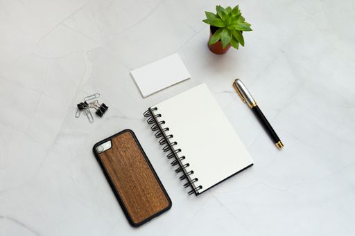 A flat lay of a blank notepad, pen, phone, clips, paper cards, and a plant on a white table