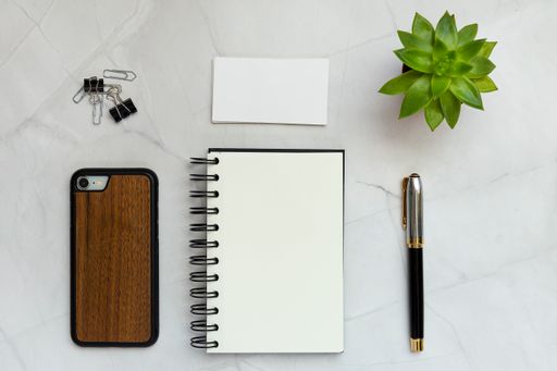 A flat lay of a blank notepad, pen, phone, clips, paper cards, and a plant on a white table