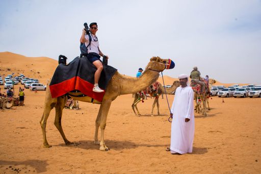 A young Arab male holding a camel with a tourist riding it in Dubai