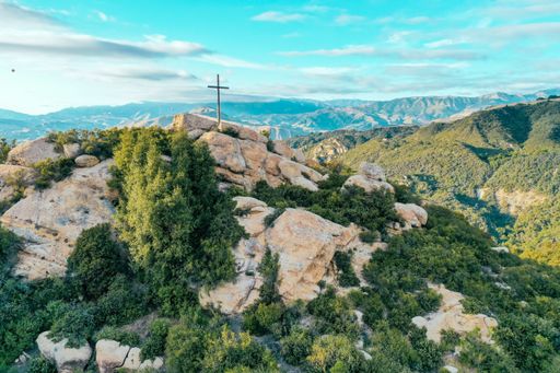 A rocky cliff covered in greenery with a cross put on the top and beautiful mountains in the background