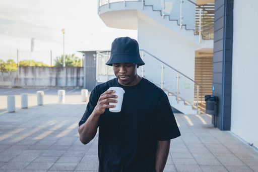 A young black man in street-style clothes holding his takeaway drink in front of a building