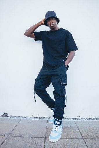 A vertical shot of a young black man in street-style clothes posing leaning on a white wall outdoors