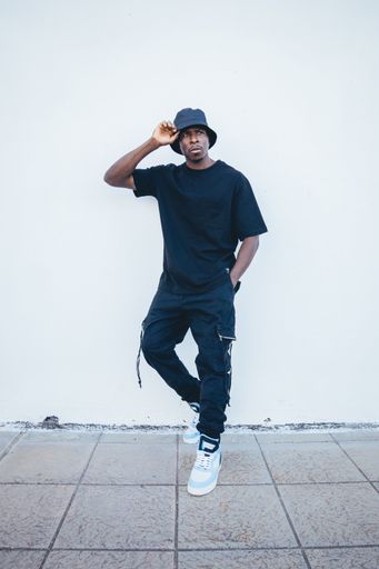 A vertical shot of a young black man in street-style clothes posing leaning on a white wall outdoors