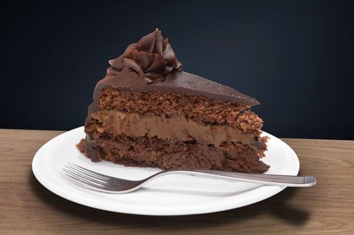A slice of rich chocolate cake with a fork on a white plate against a dark background.