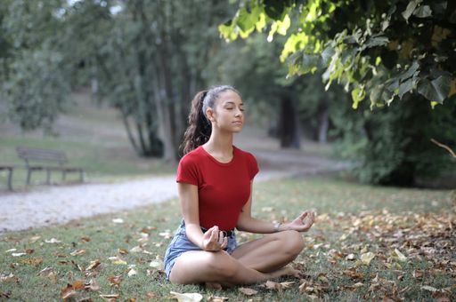 A selective shot of a female with a red shirt and denim skirt doing yoga on a green grassy field with bush and tree