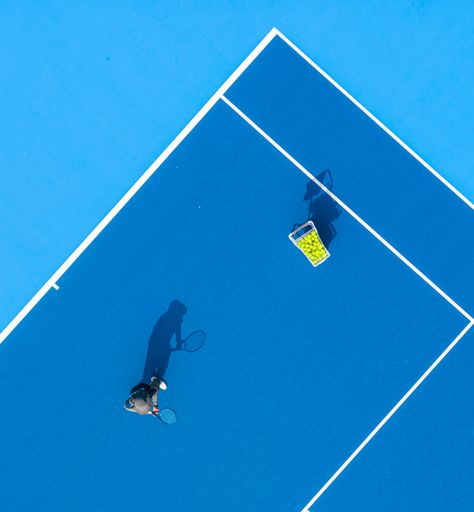 Aerial view of a tennis player practicing on a vibrant blue court with a basket of tennis balls casting a shadow.