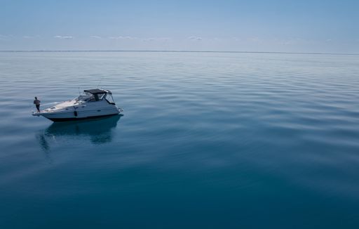 A serene scene of a lone boat on calm blue waters under a clear sky, with a person standing on deck.