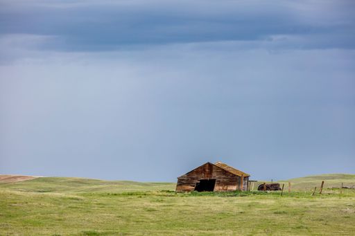 A rustic wooden barn on a vast green field under a cloudy sky in a rural landscape.