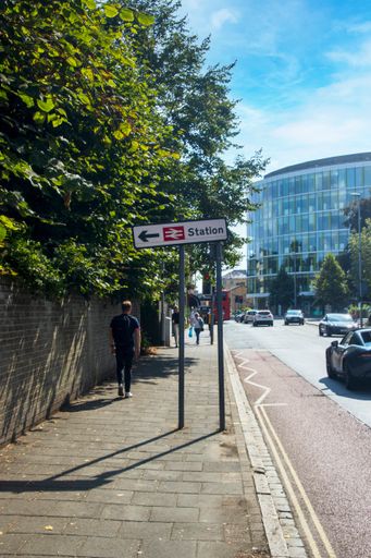 A vertical shot of a railway station sign on a footpath with pedestrians walking on a sunny afternoon