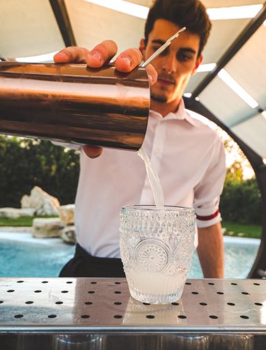 A vertical shot of a young bartender making a cocktail at the outdoor bar on the background of a pool