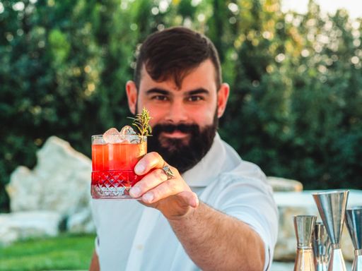 A young male with a beard showing a glass of Conte speziato cocktail standing at the outdoor bar