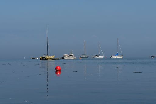Serene seascape with sailboats on calm water under a clear blue sky, featuring a prominent red buoy in the foreground.