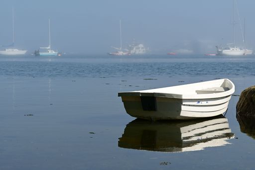 A serene image of a white rowboat on calm waters with distant sailboats shrouded in mist, capturing a peaceful coastal scene.