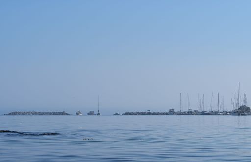 Calm ocean view with sailboats and a distant marina under a clear blue sky.