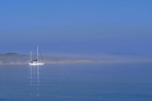 A serene sailboat on calm blue waters surrounded by mist and hills, creating a tranquil seascape.