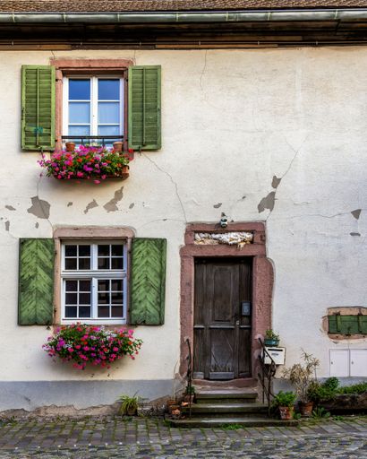 A charming rustic house facade with green shutters, colorful flowers, and weathered textures in a quaint European village.
