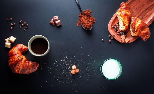 A flat lay of a breakfast setup with croissants, coffee, chocolate, and milk on a dark background.