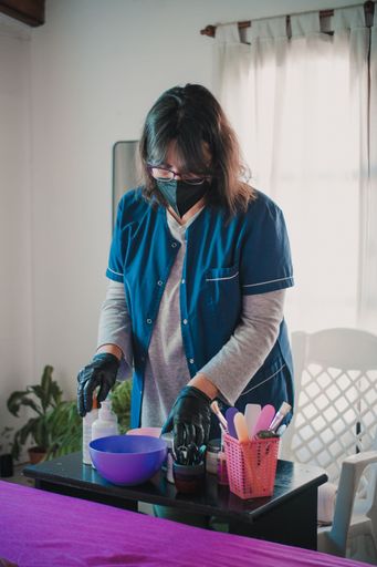 A vertical shot of a Latina beautician wearing a mask and gloves, arranging tools and bottles on a table in her studio before a treatment appointment