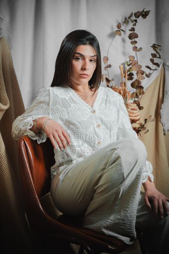 A vertical shot of a serious young Latina woman seated on a brown leather chair, modeling a white vintage lace blouse and wide-leg trousers