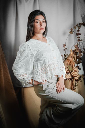A vertical shot of a fashionable young Latina woman in vintage lace blouse and gray trousers, leaning stylishly against a velvet riser in a studio